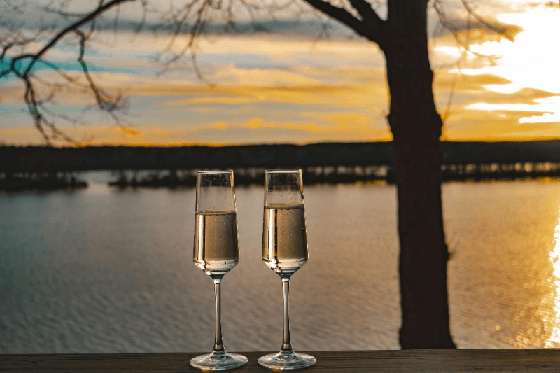 https://www.californiachampagnesabers.com/wp-content/uploads/2025/03/Two-champagne-flutes-filled-with-sparkling-wine-overlooking-a-lake-at-sunset.png